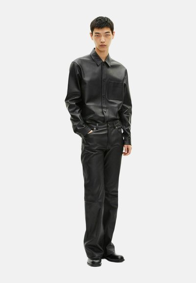 Camicia nera di pelle con maniche lunghe, design con bottoni e tasca sul petto, abbinata a pantaloni e stivali neri in pelle.