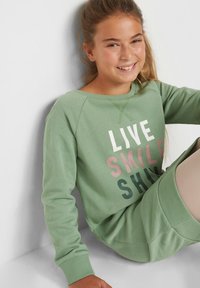 Ragazza seduta contro un muro bianco che sorride, indossando un vestito felpa verde con le parole "LIVE SMILE SHINE" sulla parte anteriore.