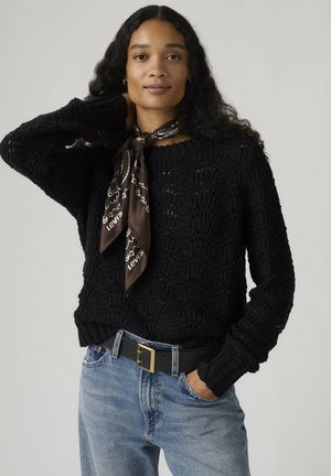Kvinde med langt, krøllet hår iført en sort strikket sweater, sort mønstret tørklæde bundet om halsen, blå jeans og sort bælte med guldlås.