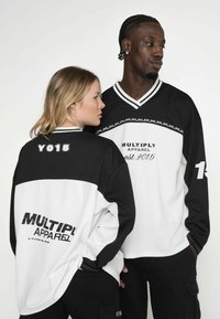 Schwarz-weiße Sporttrikots mit V-Ausschnitt, die den Text "MULTIPLY APPAREL" und Designelemente aufweisen. Auf der Rückseite steht "YO15" und "est. 2015."
