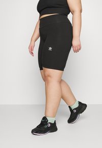 Person som bär svarta Adidas cykelshorts, svarta sneakers med pastellgröna strumpor, stående med ena benet något framåt mot enfärgad bakgrund.