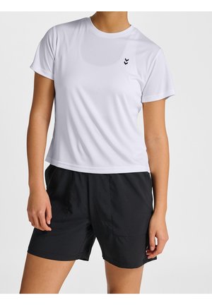 Person trägt ein weißes kurzärmeliges Sportshirt und schwarze Shorts, steht mit entspannten Armen an den Seiten vor einfarbigem Hintergrund.