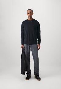 Marineblå strikkegenser med lange ermer, kombinert med grå jeans og svarte joggesko. Holder en grå veske, enkel bakgrunn.
