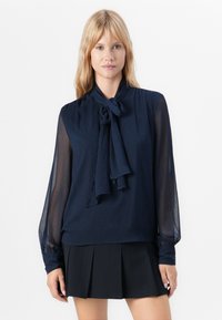 Blondinka v navy prosojni bluzici z dolgimi rokavi in zavezanim trakom okrog vratu ter črno plisirano mini krilo, stoji ob beli steni.