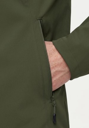 Hand in die geöffnete Tasche eines olivgrünen Jackenärmel gesteckt, zeigt Stoffstruktur und Reißverschlussdetails.