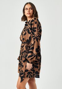 Abito camicia marrone con motivo floreale nero, dotato di maniche a sbuffo e vita cinte. Tessuto morbido con una texture leggera.