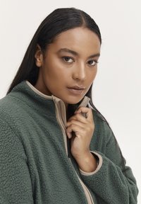 Chaqueta de forro polar verde con cremallera y detalles en beige, que presenta una superficie texturizada y un cuello alto, sostenida por un modelo con cabello largo.