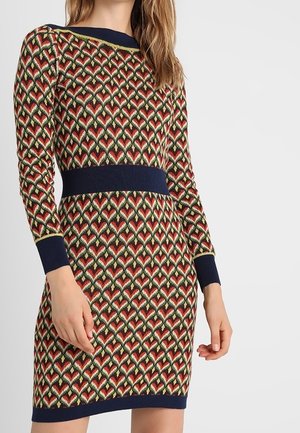 Mujer vistiendo un vestido de manga larga hasta la rodilla con un patrón geométrico en rojo, verde y blanco, y puños, cinturilla, cuello y dobladillo en azul marino.