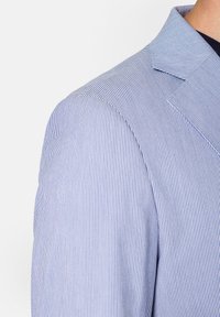 Pasiakowana kurtka w niebieskim i białym kolorze, z wyciętym kołnierzem i teksturowanym materiałem. Zbliżenie na szew ramienia oraz wzór pionowych pasków.