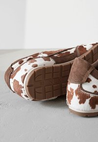 Scarpe slip-on con stampa di mucche, caratterizzate da una combinazione di suede marrone e pelliccia bianca, con suola in gomma marrone e fondo decorato.