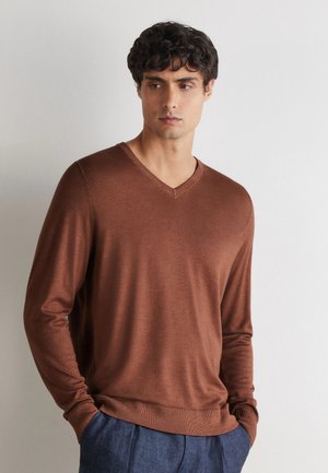 Man met kort kroezend donker haar, die een bruine lange mouwen V-hals trui en blauwe broek draagt, staat tegen een eenvoudige lichte muur.