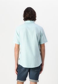 Helles mintgrünes Kurzarmhemd mit gerade geschnittenem Saum, Kragen und subtiler Textur. Getragen mit hochgerollten Denim-Shorts.
