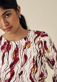 Langærmet sweater i hvid med et bølget abstrakt mønster i nuancer af rød, gul og sort. Ribbet halsudskæring og samlede skulderdetaljer.
