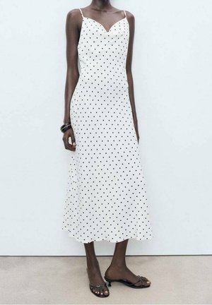 Femme portant une robe nuisette midi blanche à pois noirs, fines bretelles, bracelets noirs et sandales à talons ouvertes noires, debout sur un sol gris.
