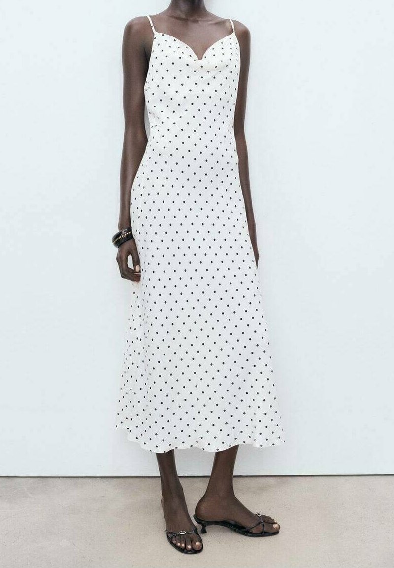 Femme portant une robe nuisette midi blanche à pois noirs, fines bretelles, bracelets noirs et sandales à talons ouvertes noires, debout sur un sol gris.