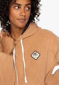 Brown fleece hoodie with a white drawstring, featuring a diamond-shaped logo patch. The fabric has a soft, textured finish.