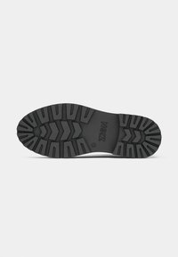 Semelle de chaussure en caoutchouc noir avec une surface texturée, présentant de profondes rainures et un emblème central, conçue pour l'adhérence et la stabilité.