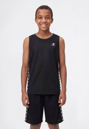 Băiat zâmbitor purtând un tricou sport negru fără mâneci Champion și pantaloni scurți asortați cu dungi cu logo pe laterale, stând în fața unui fundal alb.