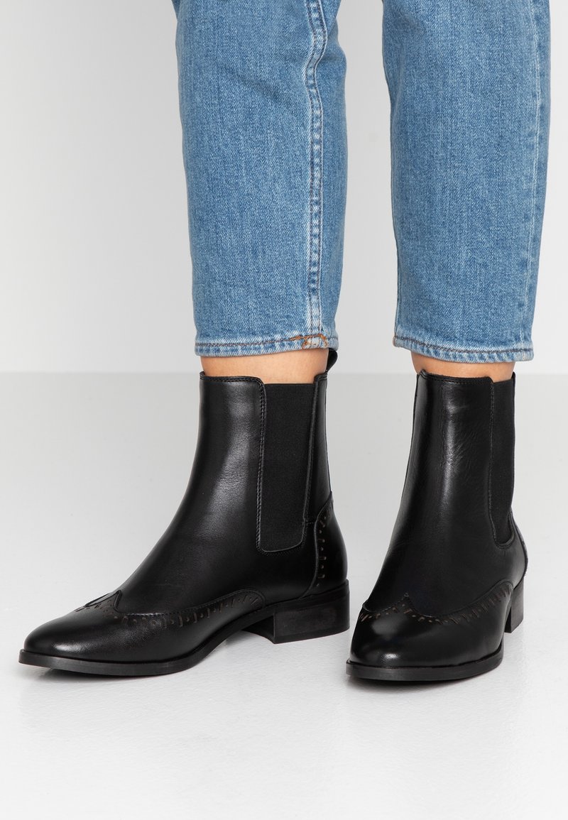 Personne portant des bottines noires en cuir avec des panneaux élastiques sur les côtés et des détails cousus, associées à un jean cropped bleu.