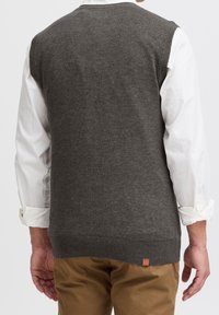 Gilet gris en tricot avec des bords côtelés, porté par-dessus une chemise blanche à boutons, arborant une petite étiquette en cuir au niveau de l'ourlet.