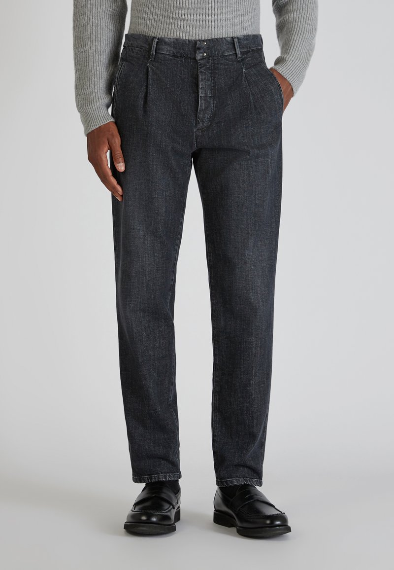Pantaloni in denim scuro con taglio dritto, dotati di chiusura con bottone, due pince sul davanti e una texture sottile. Indossati con scarpe nere.