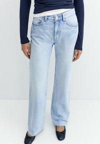 Persoană purtând blugi albaștri deschiși cu picioare largi, bluză cu mâneci lungi bleumarin și balerini negri, stând în fața unui fundal alb simplu.