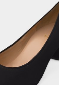 Μαύρο γυναικείο παπούτσι με χαμηλό τακούνι, στρογγυλεμένη μύτη και μπεζ εσωτερική επένδυση, εμφανιζόμενο σε κοντινή λήψη σε λευκό φόντο.