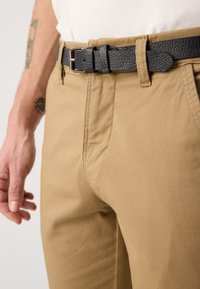 Brune chinos med en tettsittende passform, med sidelommer, og et teksturert svart lærbelte med en sølvfarget spenne.