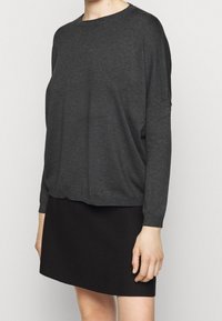 Osoba ubrana w luźny, ciemnoszary sweter z długim rękawem i dopasowaną czarną spódnicę na jednolitym jasnym tle.