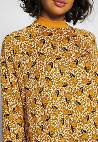 Dlouhý rukáv žlutého topu s květinovým vzorem s černými obrysy zobrazenými na postavách. Materiál vypadá hladce a má vysoký límec.