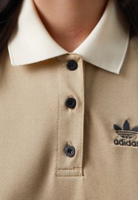 Beige polo tričko s kontrastním krémovým límečkem, třemi černými knoflíky a vyšitým logem Adidas v tmavě modré barvě.