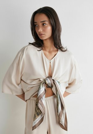 Kobieta o długich ciemnych włosach, ubrana w beżowy luźny top zawiązany wzorzystym szalikiem i dopasowane beżowe spodnie, stojąca na tle jednolitej ściany.