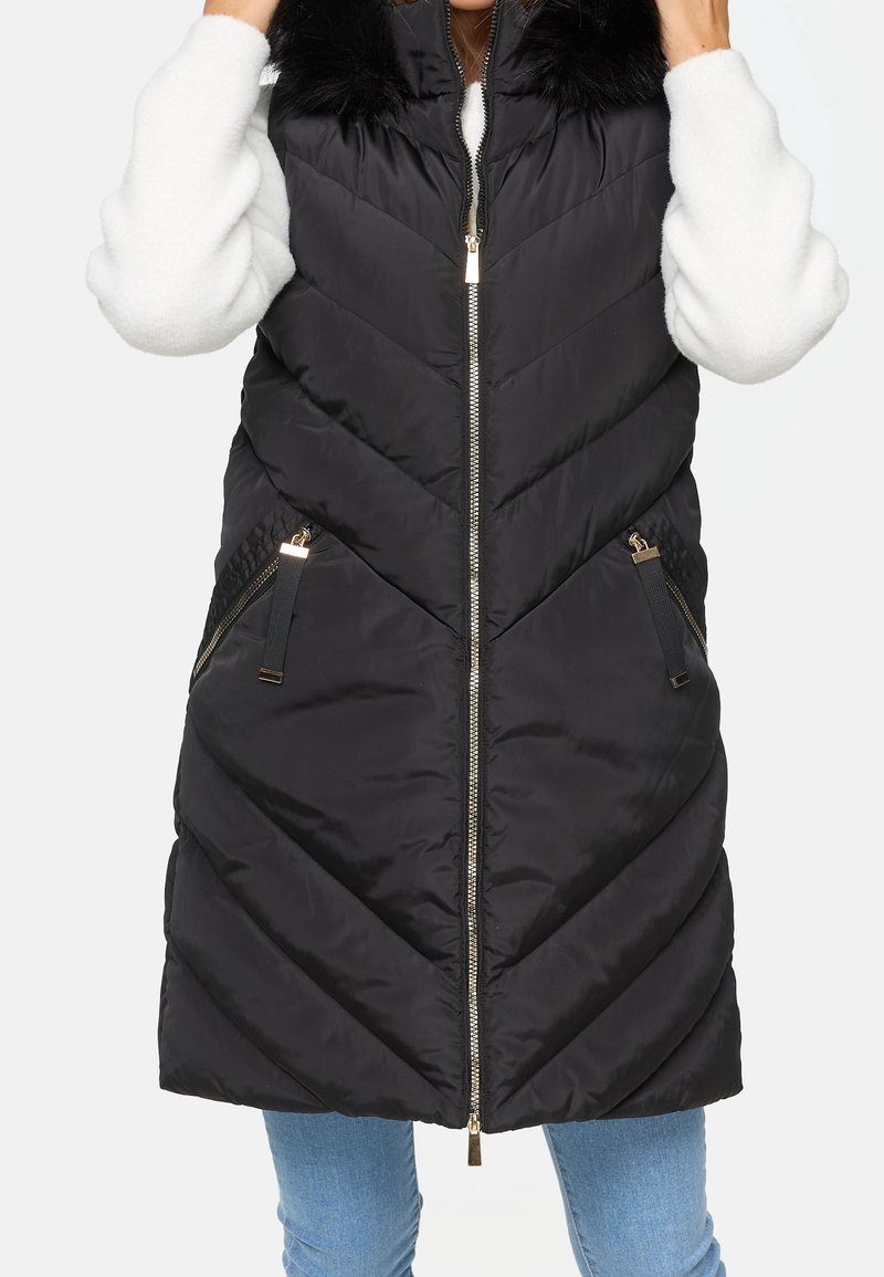 Veste sans manches noire matelassée avec motif chevron, capuche doublée de fourrure, fermeture éclair dorée et poches latérales zippées, portée avec un pull blanc et un jean bleu.