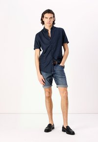 Chemise à manches courtes bleu marine avec des pois blancs, associée à un short en denim retroussé et des chaussures noires à enfiler, illustrant une tenue d'été décontractée.