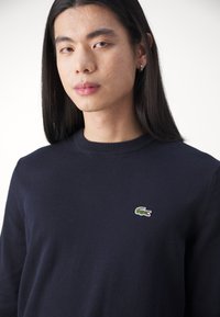 Pull marine en tissu doux, avec un col rond et des poignets côtelés, arborant un petit logo de crocodile vert sur la poitrine gauche.