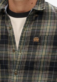 Camicia a quadri in verde scuro, blu navy e giallo. Presenta bottoni marroni, taschino sul petto con etichetta del marchio e tessuto morbido.