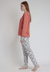 Mujer de perfil, descalza, vistiendo una blusa de manga larga color coral y pantalones de pijama blancos con un patrón de caras de animales de dibujos animados sobre un fondo claro.