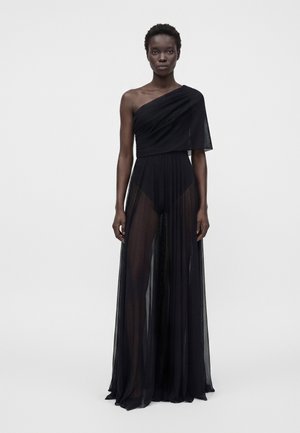 Kvinde iført en sort one-shoulder kjole med gennemsigtig plisseret nederdel og tætsiddende bodice, stående mod en ensfarvet lys baggrund.