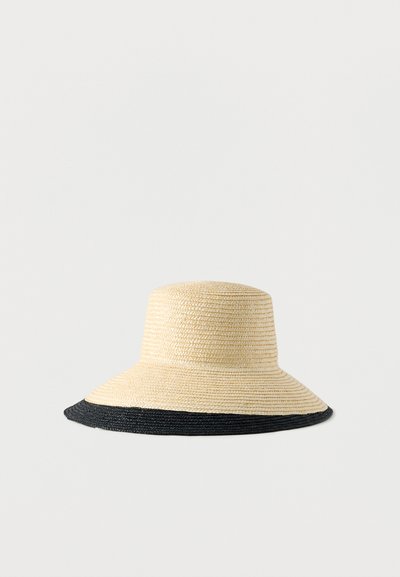 Καπέλο με φαρδύ γείσο από άχυρο σε φυσικό χρώμα, με μαύρο υφαντό διακοσμητικό κατά μήκος της άκρης. Κυκλικό σχήμα, με υφή στην επιφάνεια.