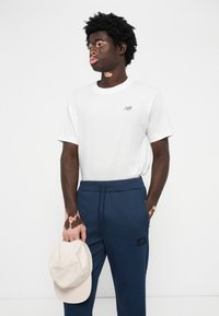 Tricou alb cu guler rotund, având un mic logo gri, asortat cu pantaloni jogger bleumarin și o șapcă bej ținută în mână. Material neted, design casual.