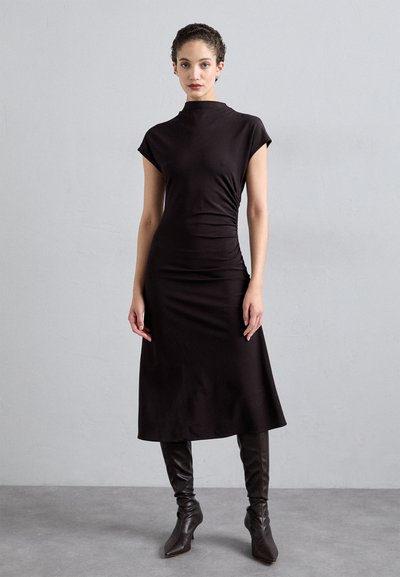 Robe noire ajustée avec un col montant, des manches courtes et un détail froncé sur le côté, associée à des bottes pointues marron montantes jusqu'aux genoux.