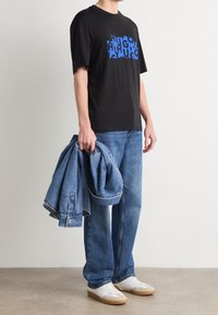Camiseta negra con estampado de logo azul, chaqueta de mezclilla azul clara sostenida en la mano, jeans azul claro y zapatillas blancas con detalles en color beige.