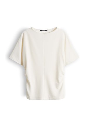 Blouse à manches courtes couleur crème avec un col rond et des détails subtils de fronces sur les côtés, présentée sur un fond blanc.