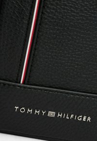 Billetera de cuero negro texturizado con un logo plateado, rayas de acento rojas y blancas, y líneas limpias. El hardware notable añade detalle.