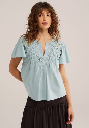 Žena s kučeravými hnedými vlasmi nosí svetlomodrú vyšívanú blúzku s krátkymi rukávmi a čiernu sukňu, stojaca pred jednoduchým pozadím.