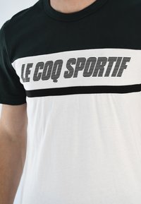 Camiseta corta de algodón en blanco y negro, con un diseño a rayas y el texto "LE COQ SPORTIF" en letras negras y en negrita a través del pecho.