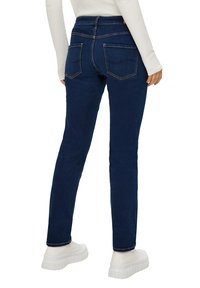 Un jean skinny en denim bleu foncé avec une taille haute, des poches arrière et une texture lisse. Associé à des baskets à plateforme blanches.