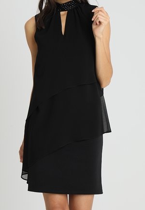 Woman in sleeveless black dress with layered asymmetrical hem and beaded high neckline, standing against a plain light background.