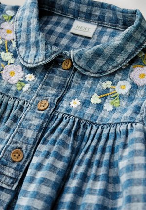 Chemise en denim avec un motif à carreaux bleu et blanc, des fleurs brodées, des boutons en bois et un col arrondi avec des détails de pinces.