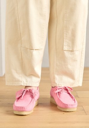 Roza nubuk mokasinke s čipkami, z teksturirano površino in svetlo bež gumijastim podplatom, v kombinaciji s bež hlačami.